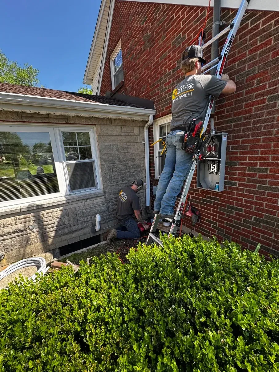 Worker on a ladder repairing a red brick wall beside a house with a hedge below