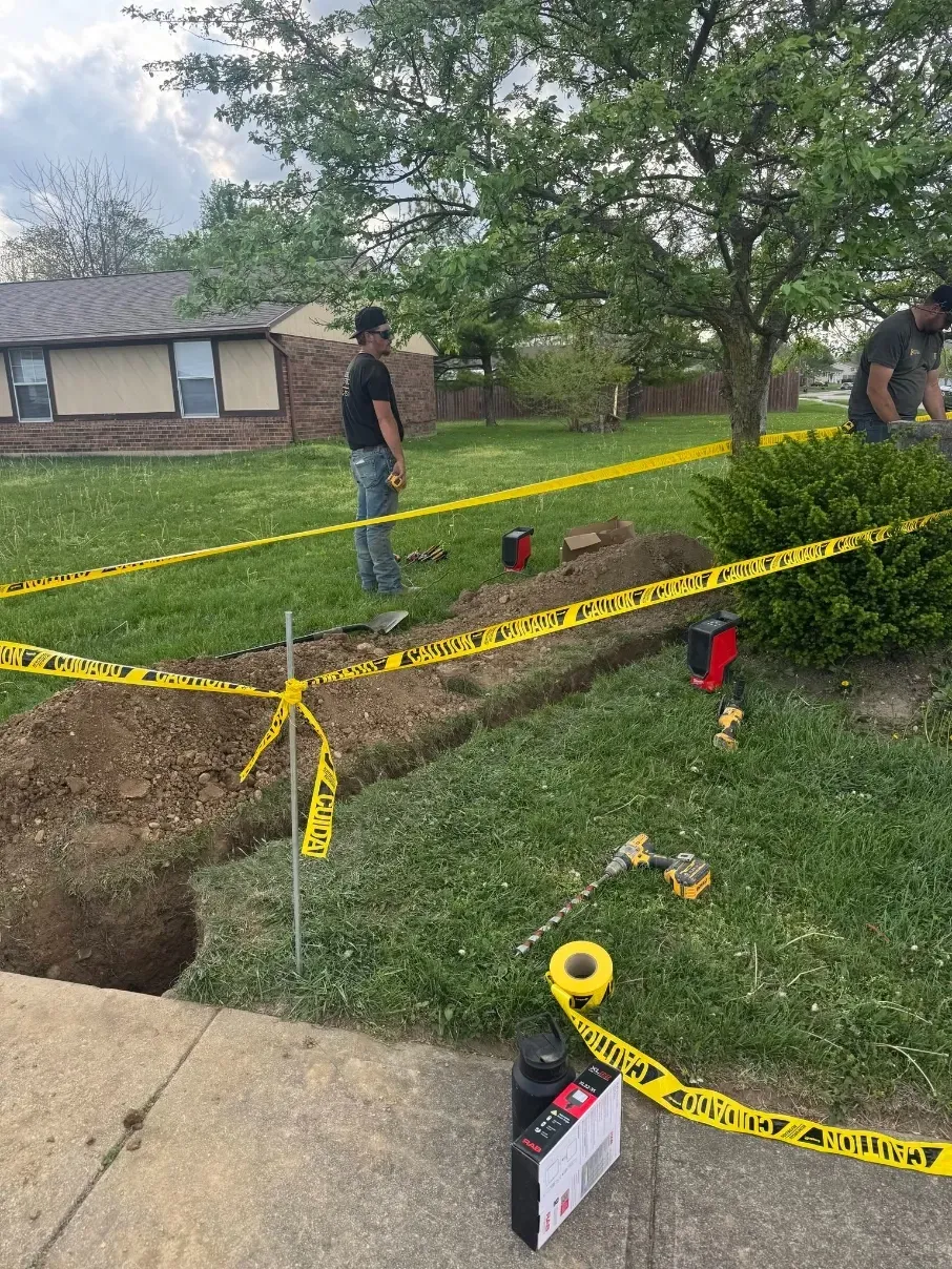 Utility crew inspecting a yard trench marked with yellow caution tape and tools near a house