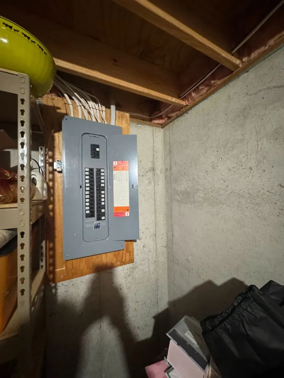 Basement wall with an electrical breaker panel mounted on wood framing under exposed joists.