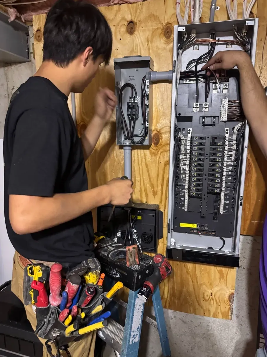 Technician wiring an electrical breaker panel in a workshop, with tools on a belt.