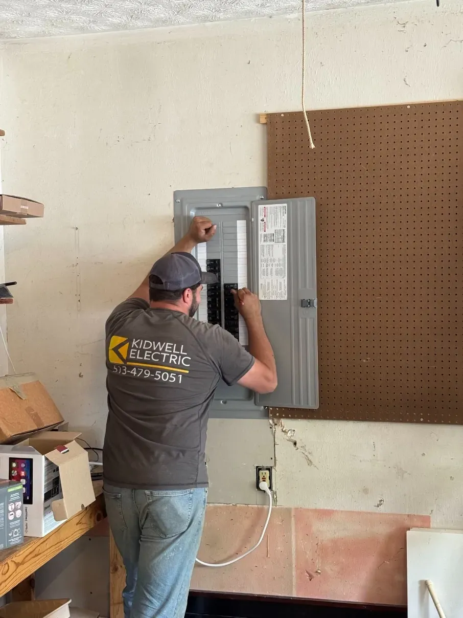 Electrician inspecting a gray breaker panel beside a pegboard wall in a garage.