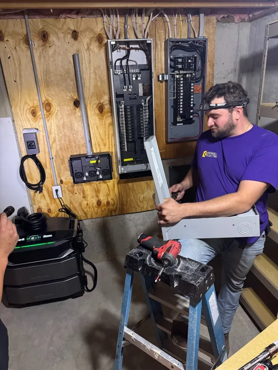 Man in purple shirt inspecting an electrical panel in a workshop, holding a long component.