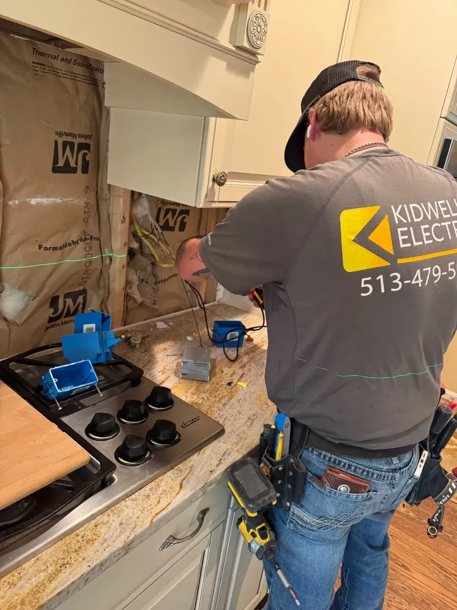 Electrician working on kitchen wiring behind a stove, with exposed wall and tools on the counter.