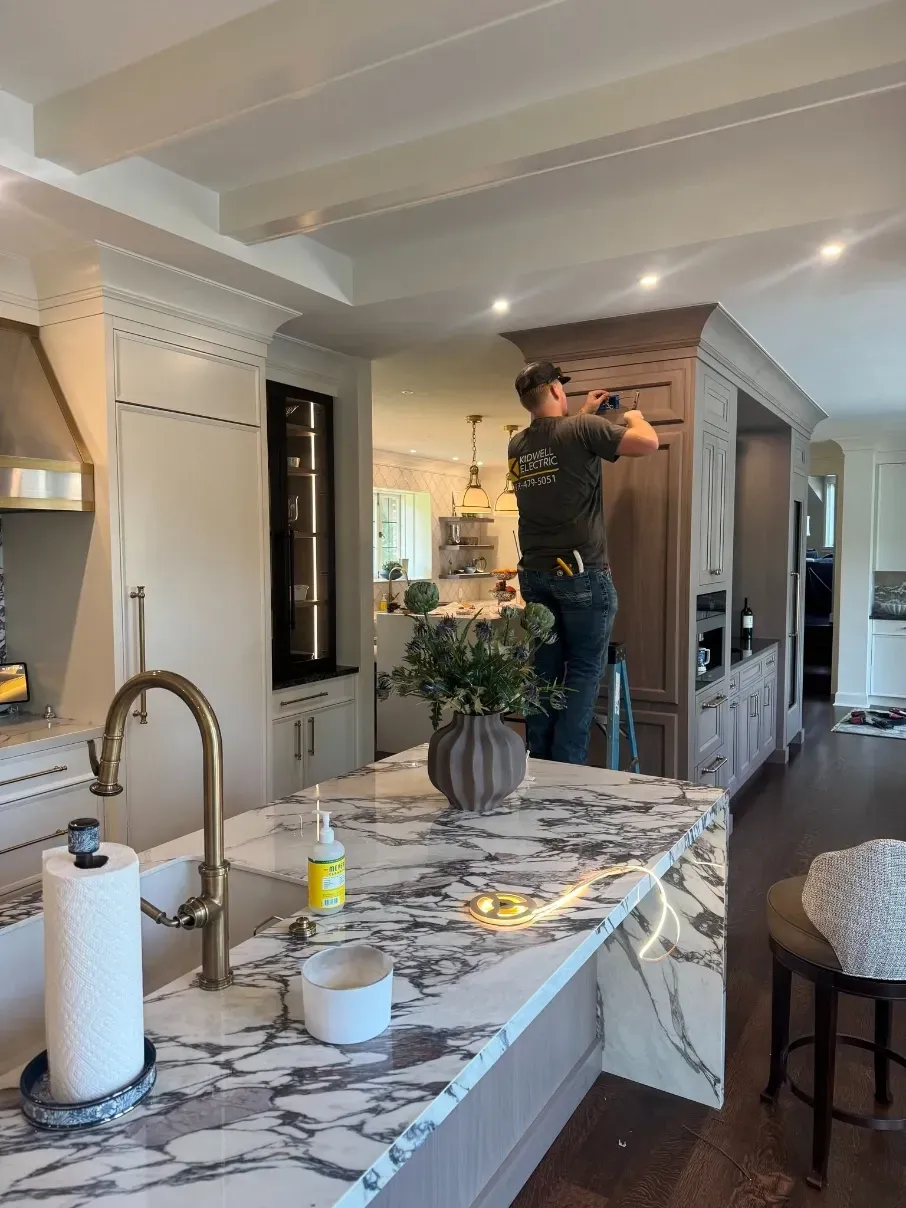 Worker installing a ceiling light in a modern kitchen with a marble island and sink.