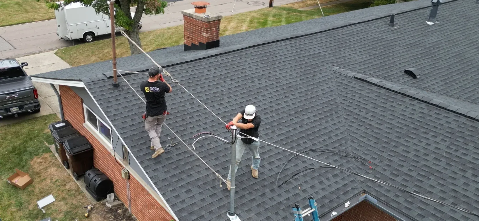 Two roof workers pulling a rope across a shingled rooftop near a brick house.