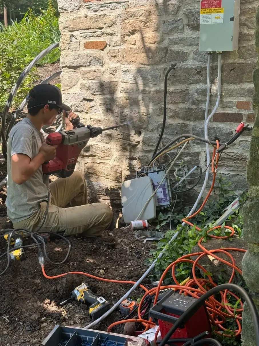 Worker using power tools beside a stone wall, surrounded by cords and equipment outdoors