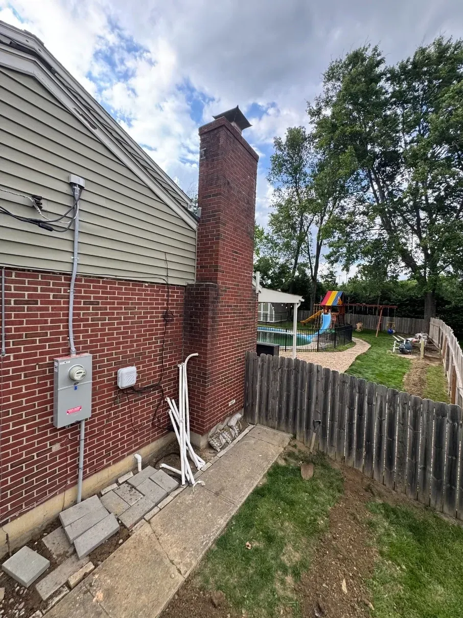 Side of brick house with chimney, utility meters, and fenced backyard with trees and a small play area.