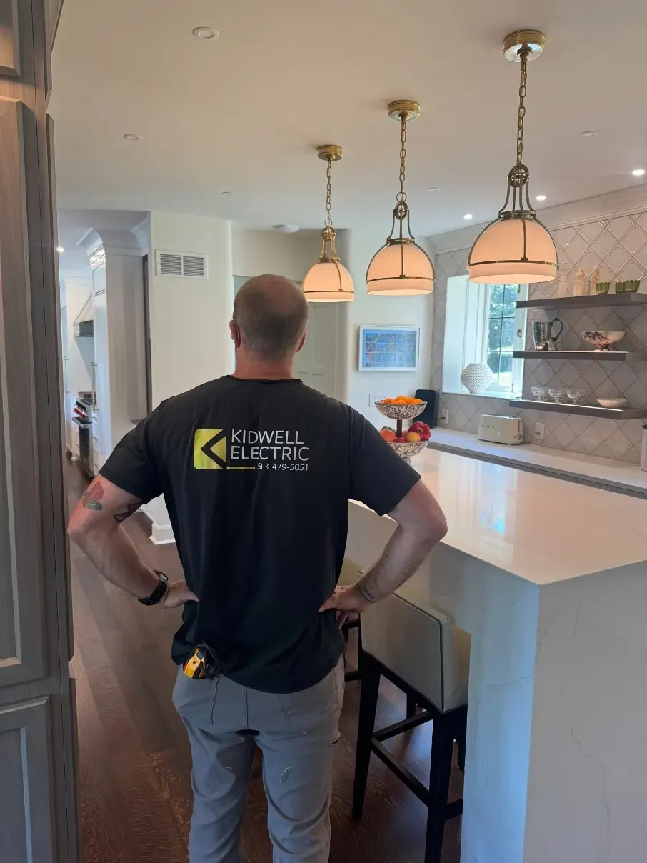Electrician standing in a bright kitchen under pendant lights, inspecting a countertop and island stools.