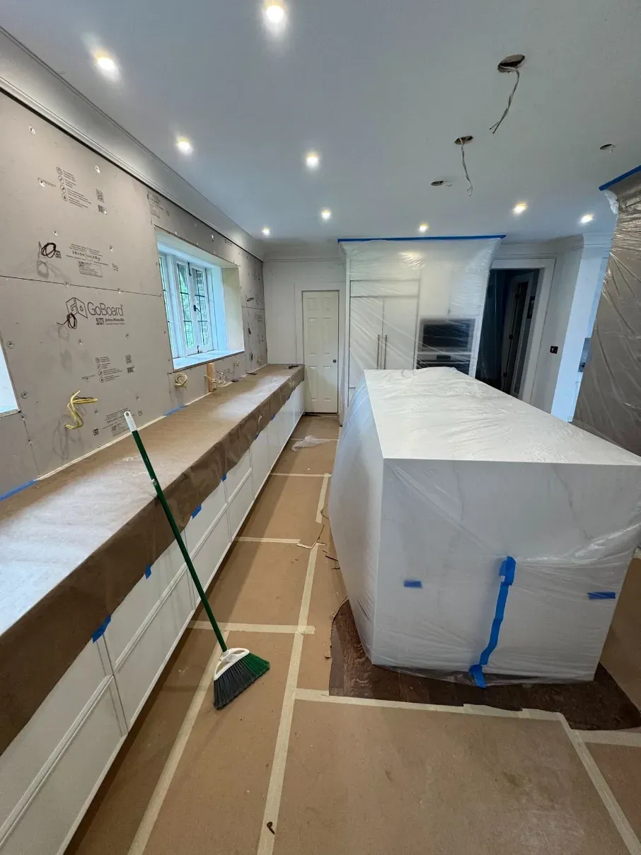 Kitchen under renovation with white island wrapped in plastic, cardboard floor protection, and workbench along wall