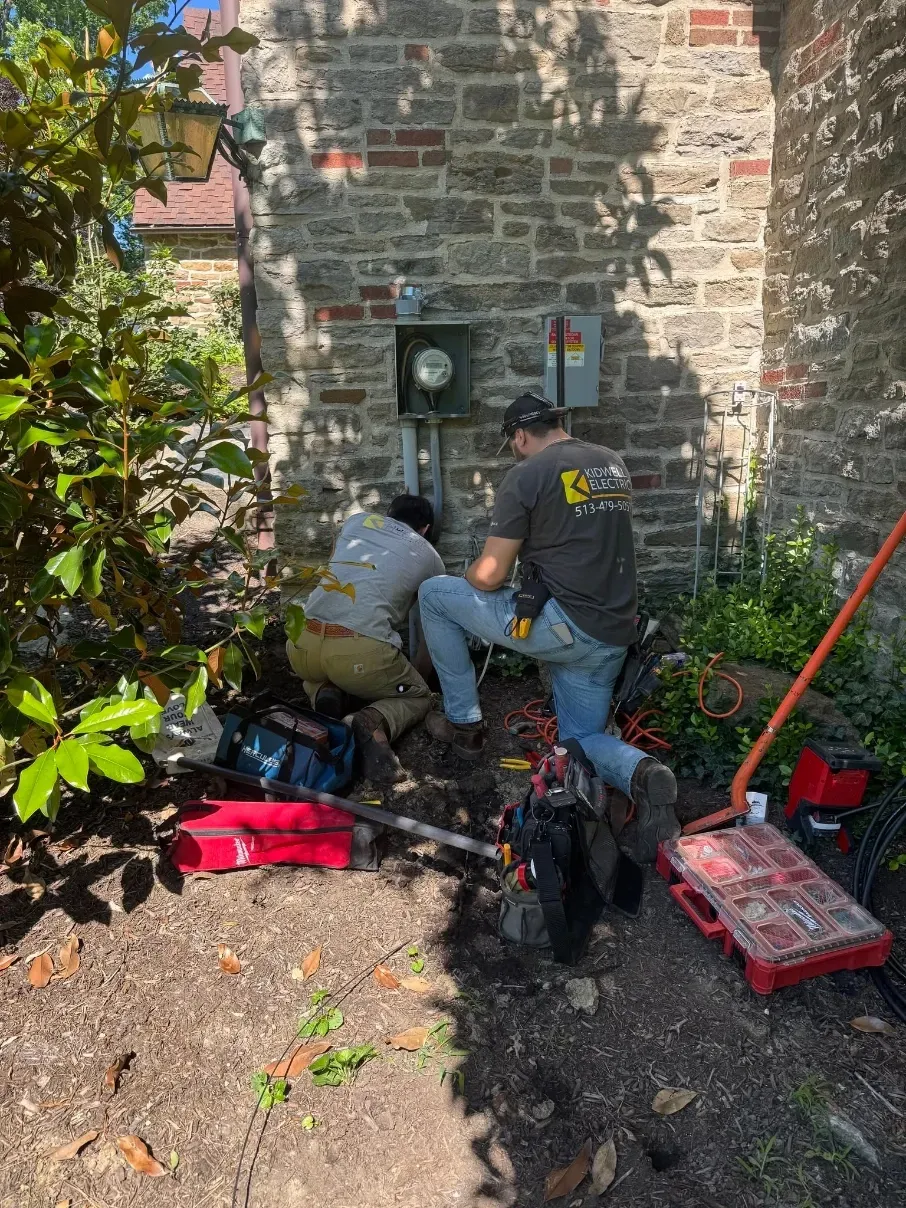 Two workers crouch by a stone wall, repairing an electrical box with tools and a red tool case nearby.