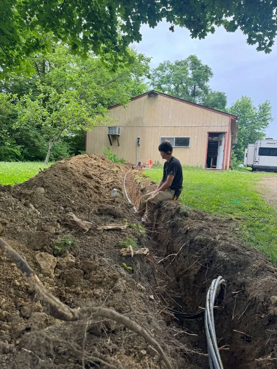 Worker digging a trench beside a large dirt mound near a tan shed in a grassy yard