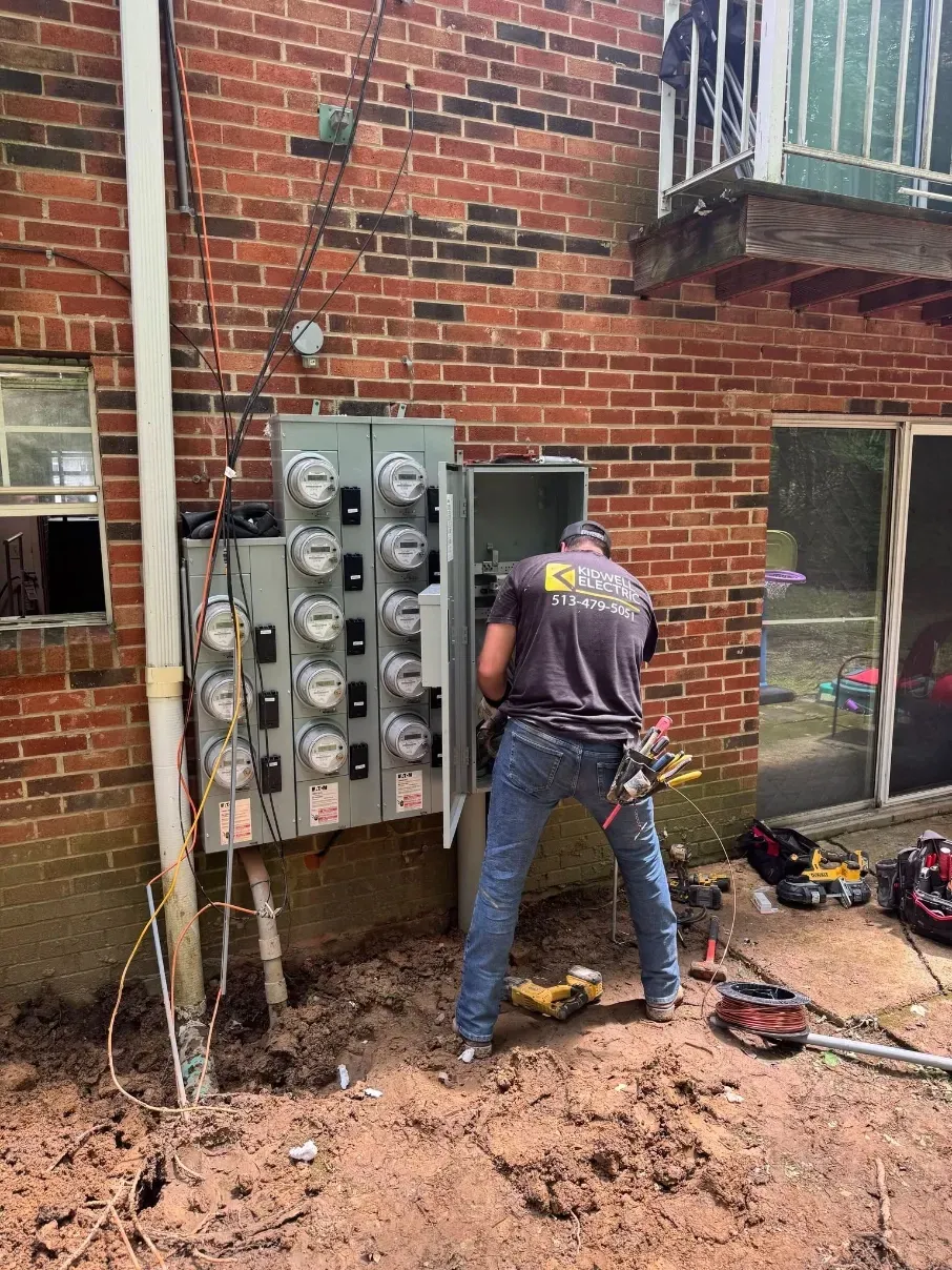 Worker repairs electrical panels on a brick building exterior, standing in muddy ground.