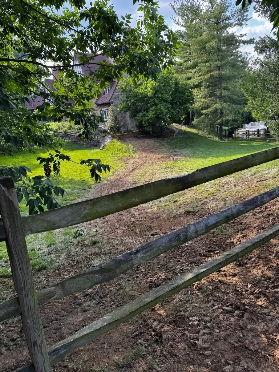 Wooden fence beside a grassy path leading to a house under leafy trees on a sunny day