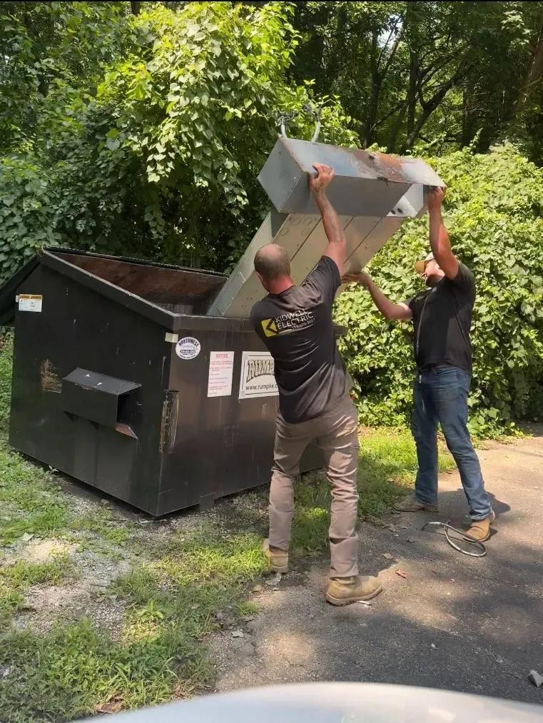 Two workers lift a metal dumpster lid outdoors beside a black dumpster in a wooded area.