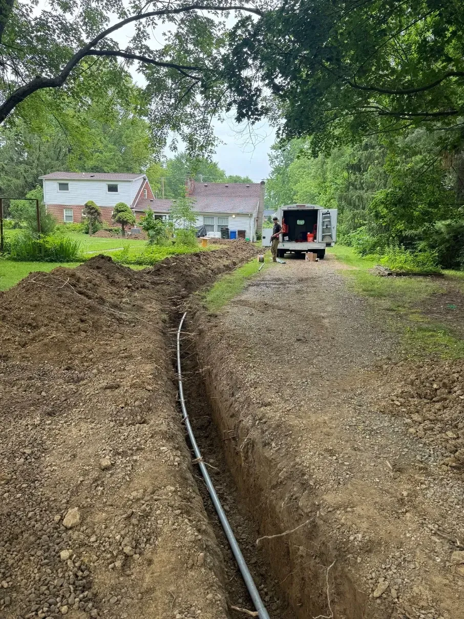 Trenched dirt driveway with a black pipe, leading to a work truck near houses and trees.