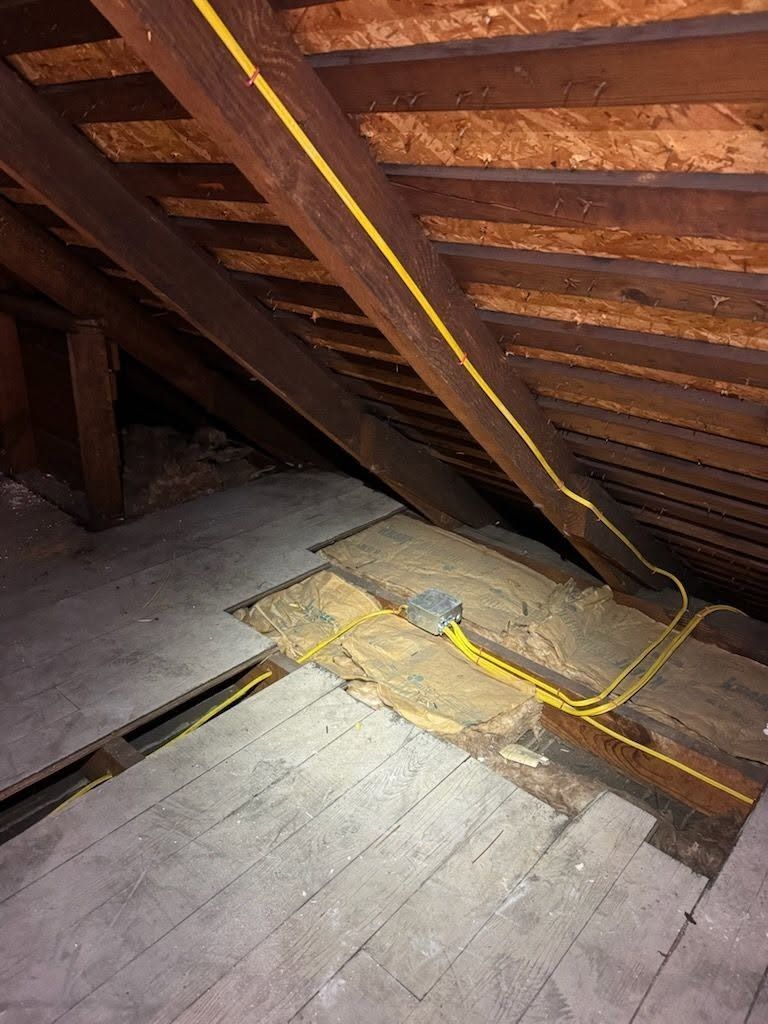 Attic floor with exposed wooden beams, insulation, and yellow electrical wiring along the rafters.