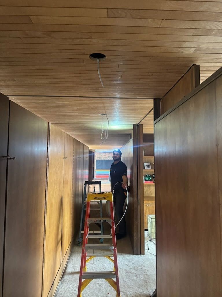Person standing on a ladder in a narrow hallway with wooden walls and ceiling under renovation