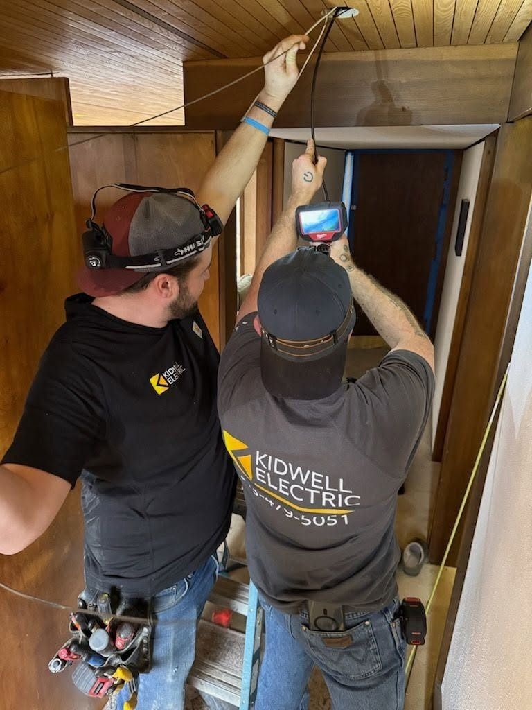 Two electricians working on ceiling wiring in a wood-paneled hallway, one holding a tool and the other a device.