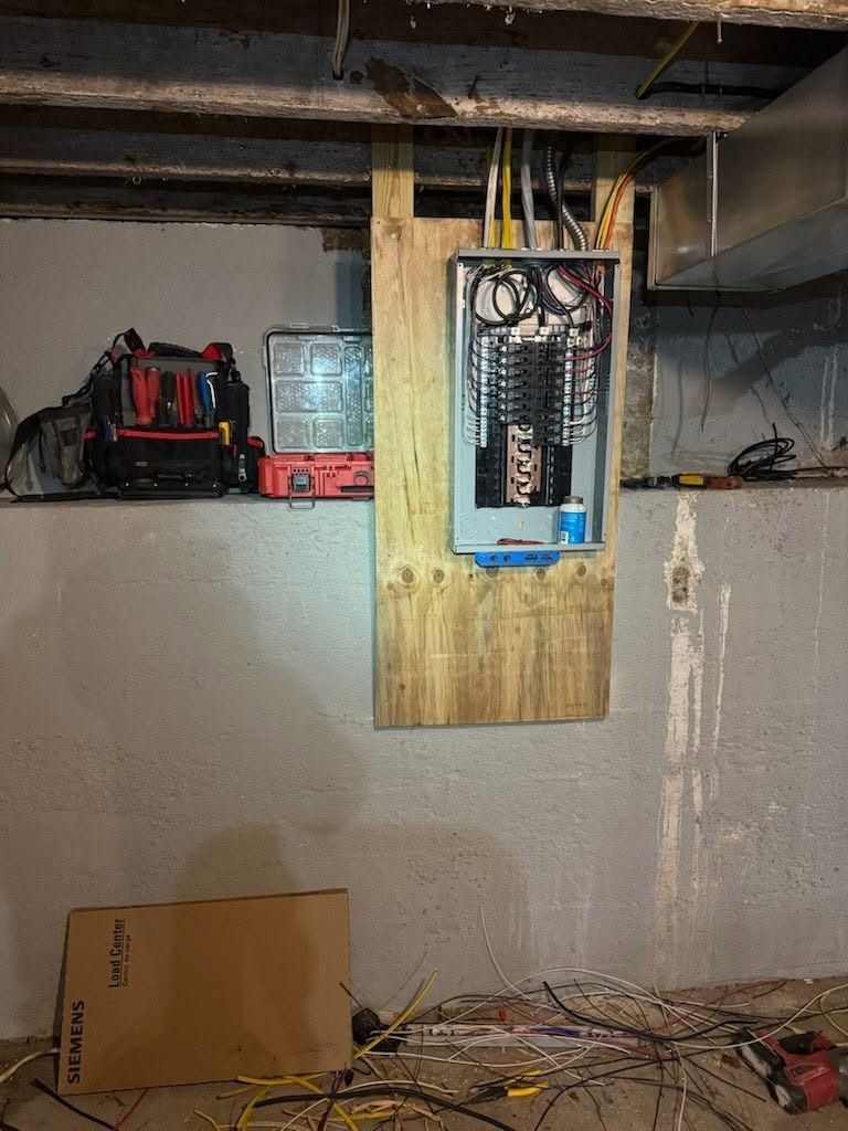 Basement wall with an open electrical panel on plywood, tools and clutter nearby.