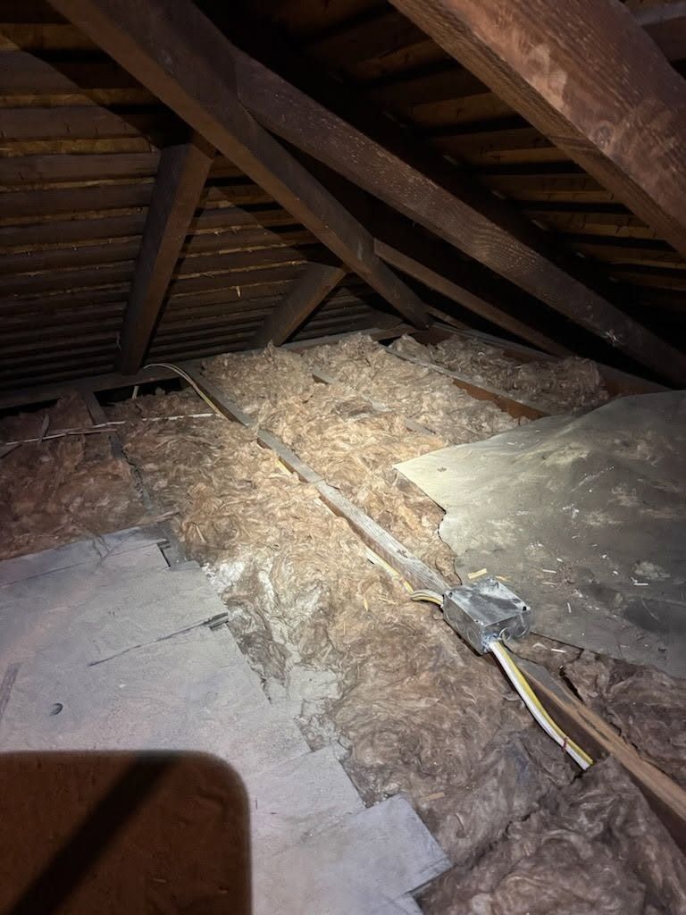 Attic with exposed wooden rafters, insulation debris, and a damaged floor area under dim light