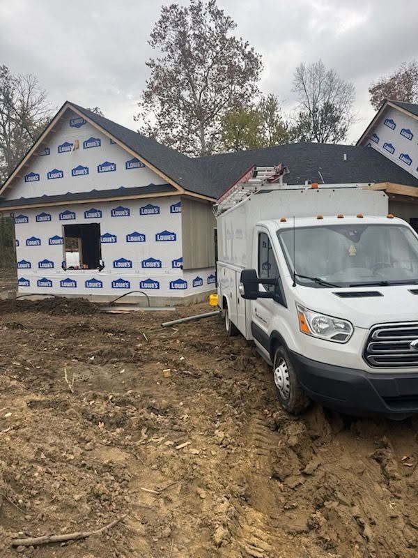 White van beside a house under construction, wrapped in housewrap, on a muddy dirt lot.