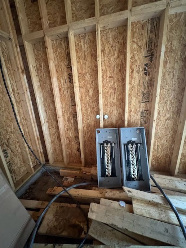 Interior of a small unfinished room with exposed wooden studs, open breaker panels, and construction debris on the floor