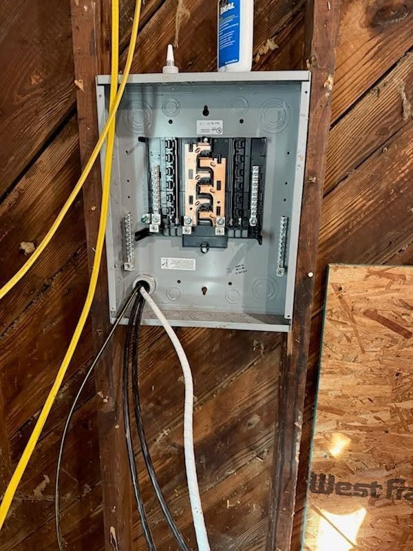 Open electrical breaker panel mounted on unfinished wooden wall with yellow and white cables nearby