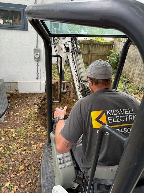 Worker in gray cap operating a compact lift in a yard beside a house.
