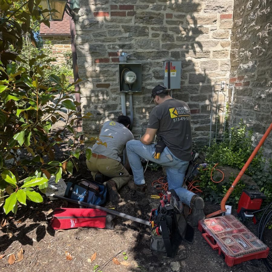 Two workers kneel by a stone wall, inspecting utility meters and tools in a small outdoor area.