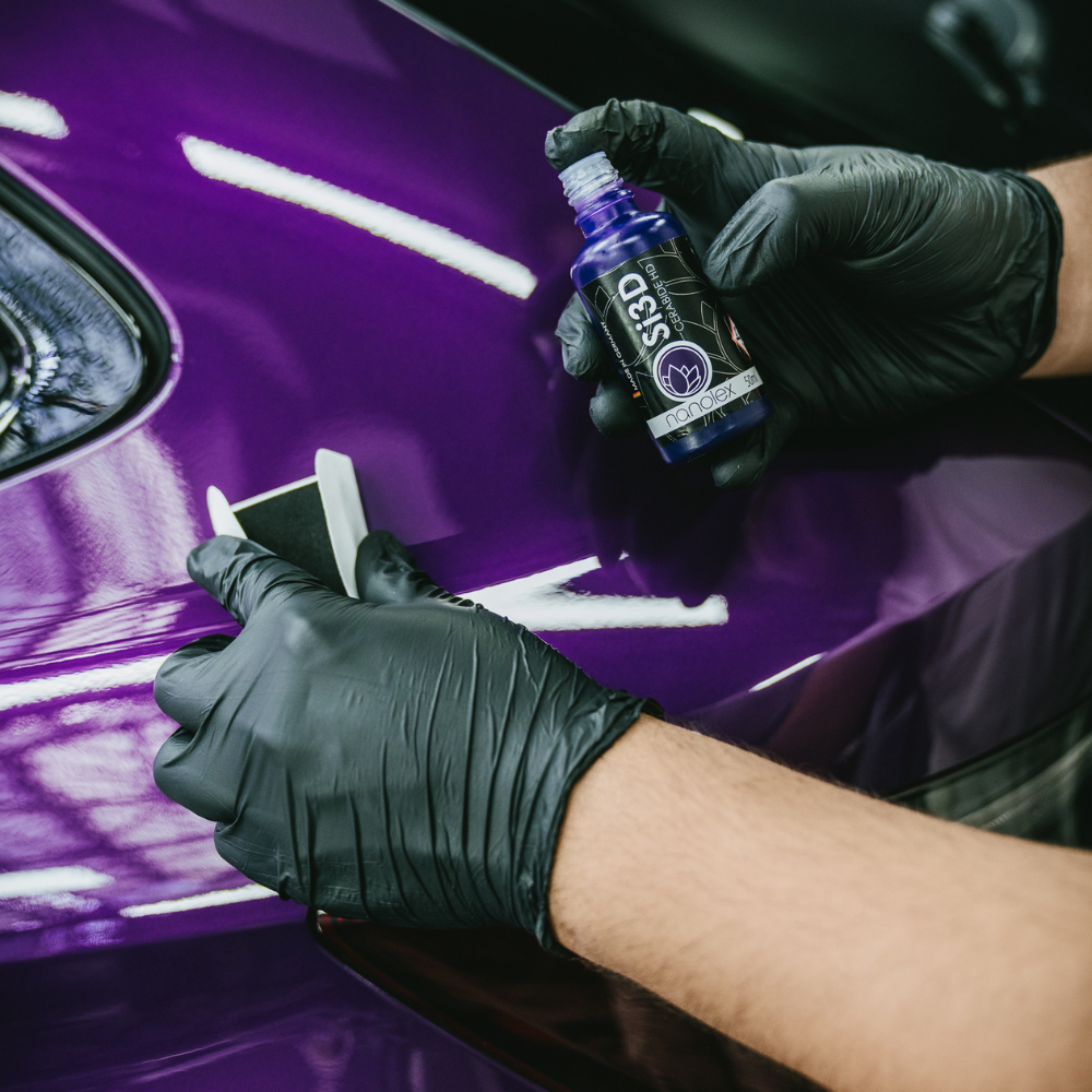 Des mains gantées appliquent du produit de polissage sur un panneau de carrosserie violet à l'aide d'un flacon de detailing.