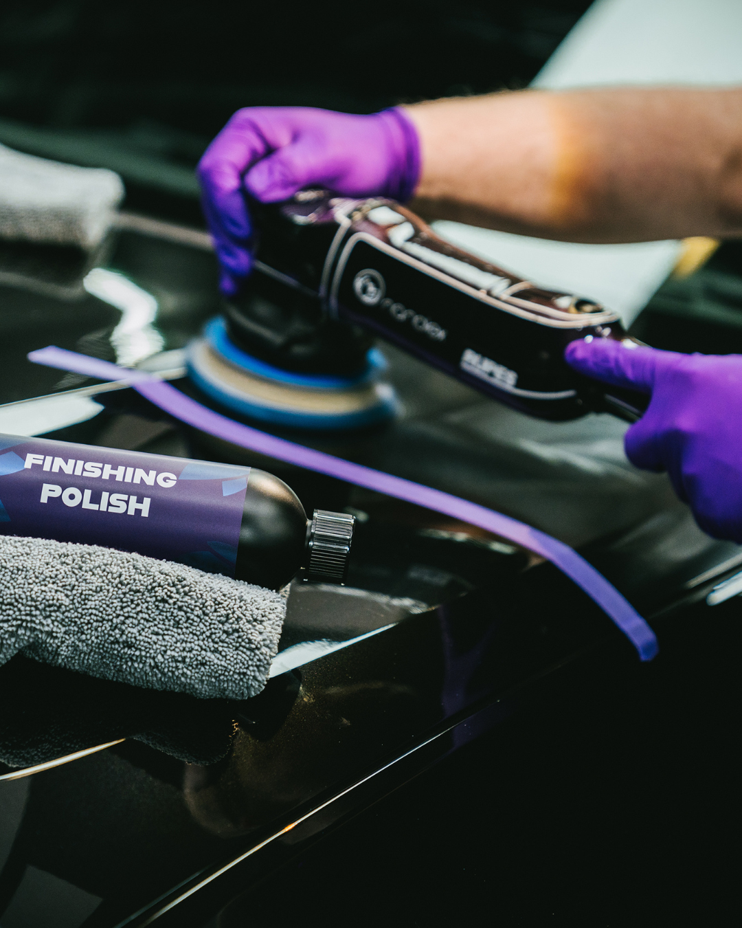 Des mains gantées de violet polissent une pièce automobile noire avec une polisseuse portative sur un établi.
