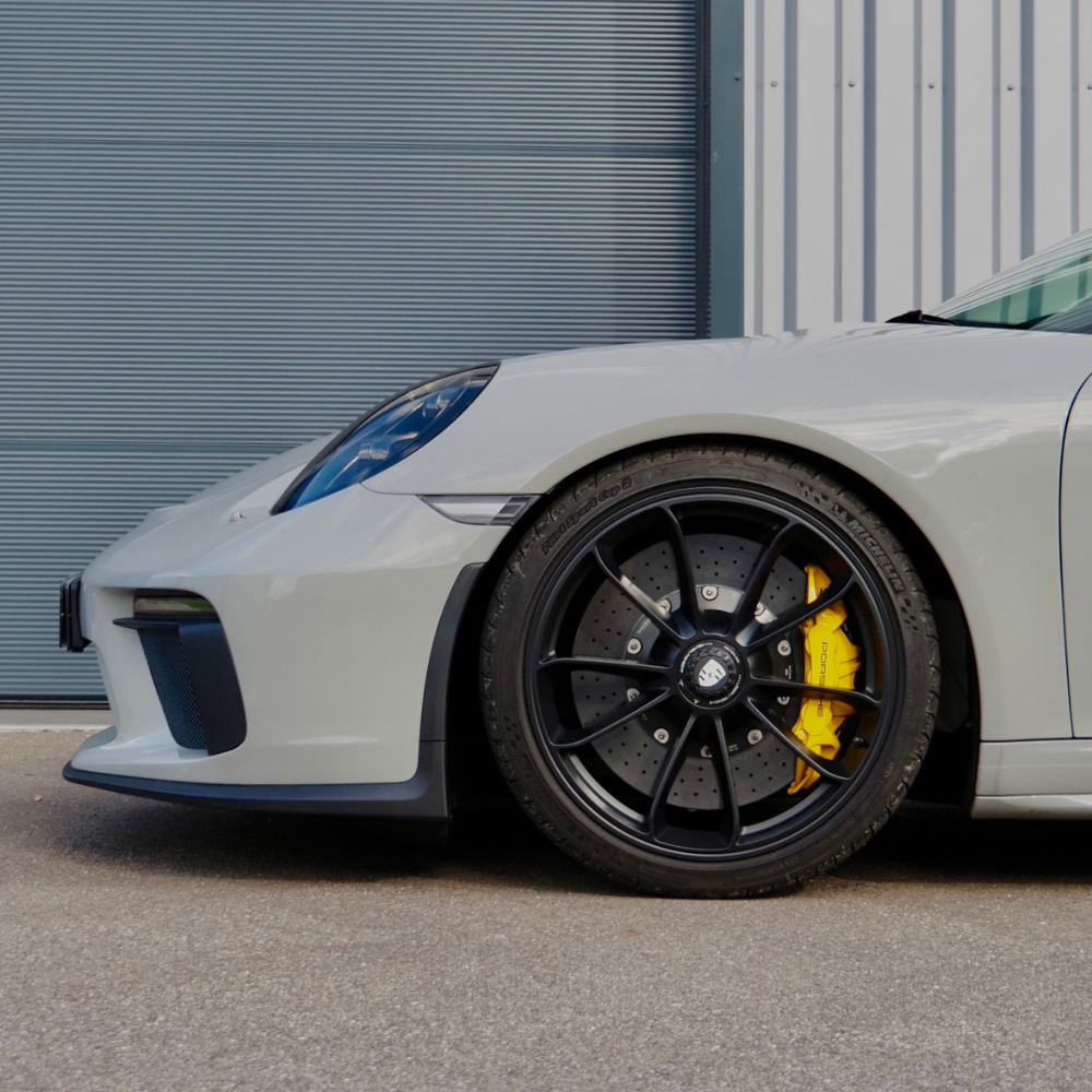 Voiture de sport blanche avec jantes noires et étriers de freins jaunes garée près d'une porte de garage métallique