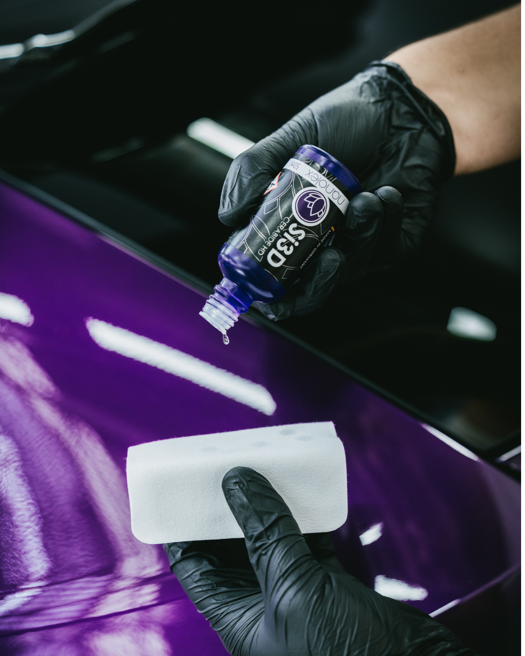 Des mains gantées appliquent du produit de polissage sur la surface violette d'une voiture à l'aide d'un applicateur blanc.