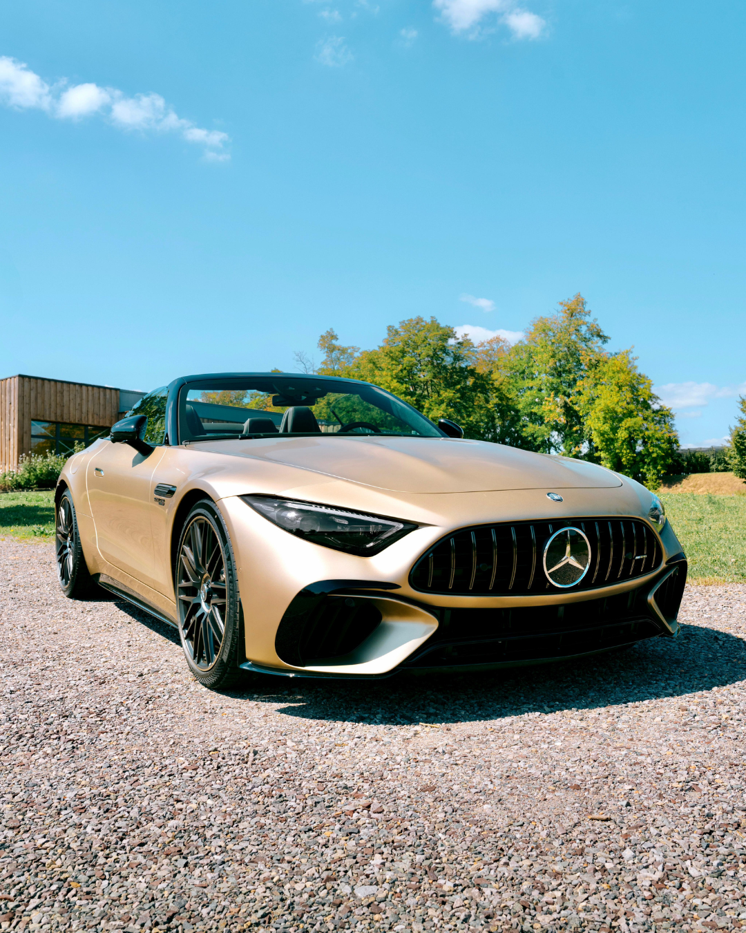 Une Mercedes-Benz décapotable beige est garée sur du gravier sous un ciel bleu, avec des arbres et une clôture en bois à l'arrière-plan.