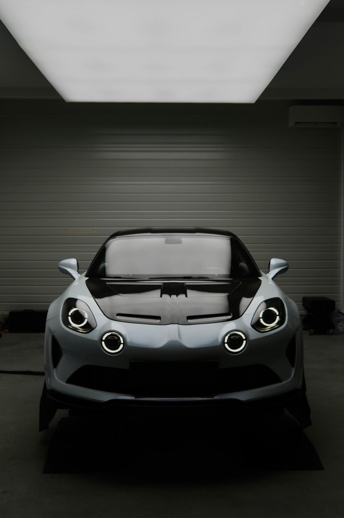 Vue de face d'une voiture de sport argentée dans un garage sombre, éclairé par un panneau lumineux au plafond.