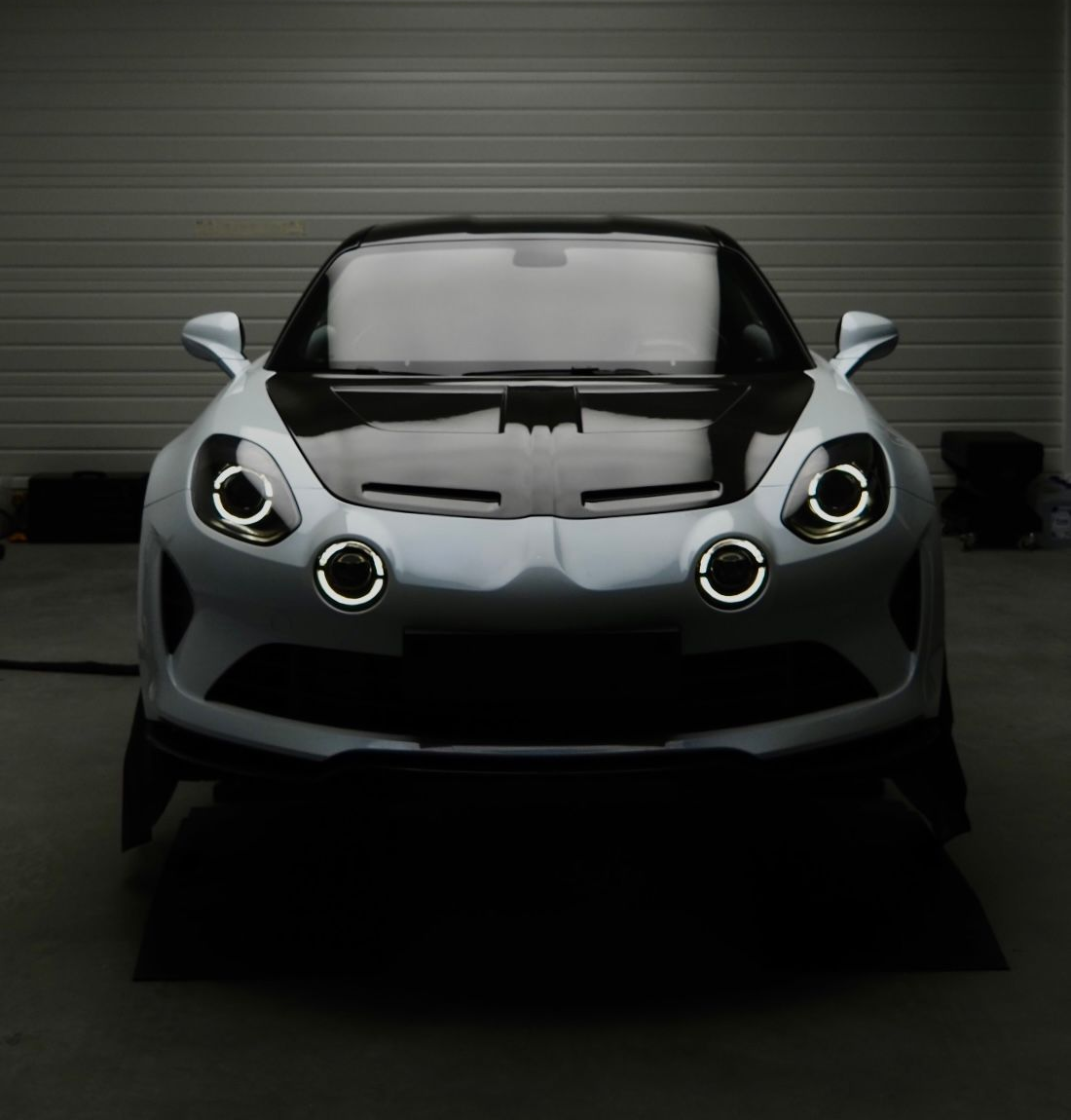 Vue de face d'une voiture de sport argentée aux phares ronds lumineux dans un garage faiblement éclairé