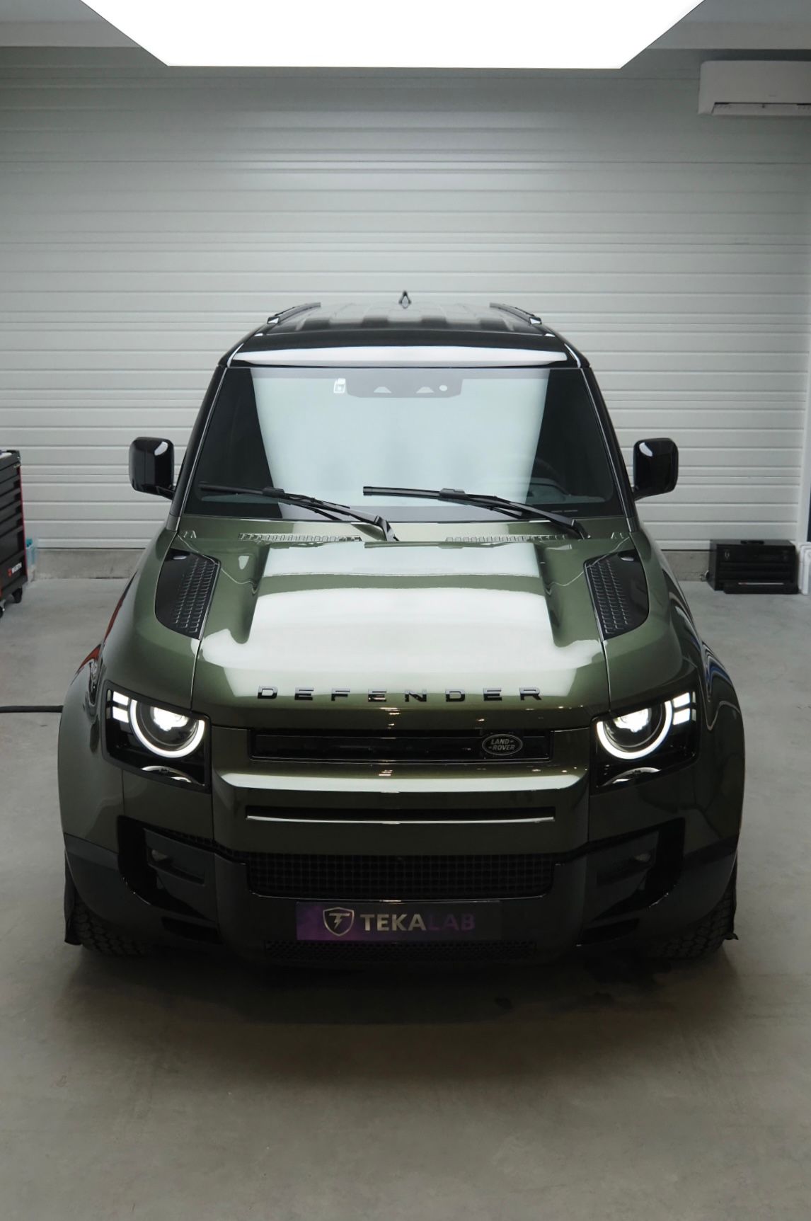 SUV vert garé à l'intérieur, adossé à une porte de garage blanche, vue de face avec phares allumés