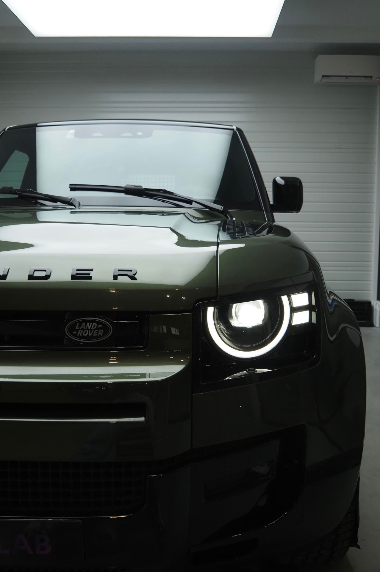 Vue de face d'un Land Rover vert dans un garage, avec un phare allumé.
