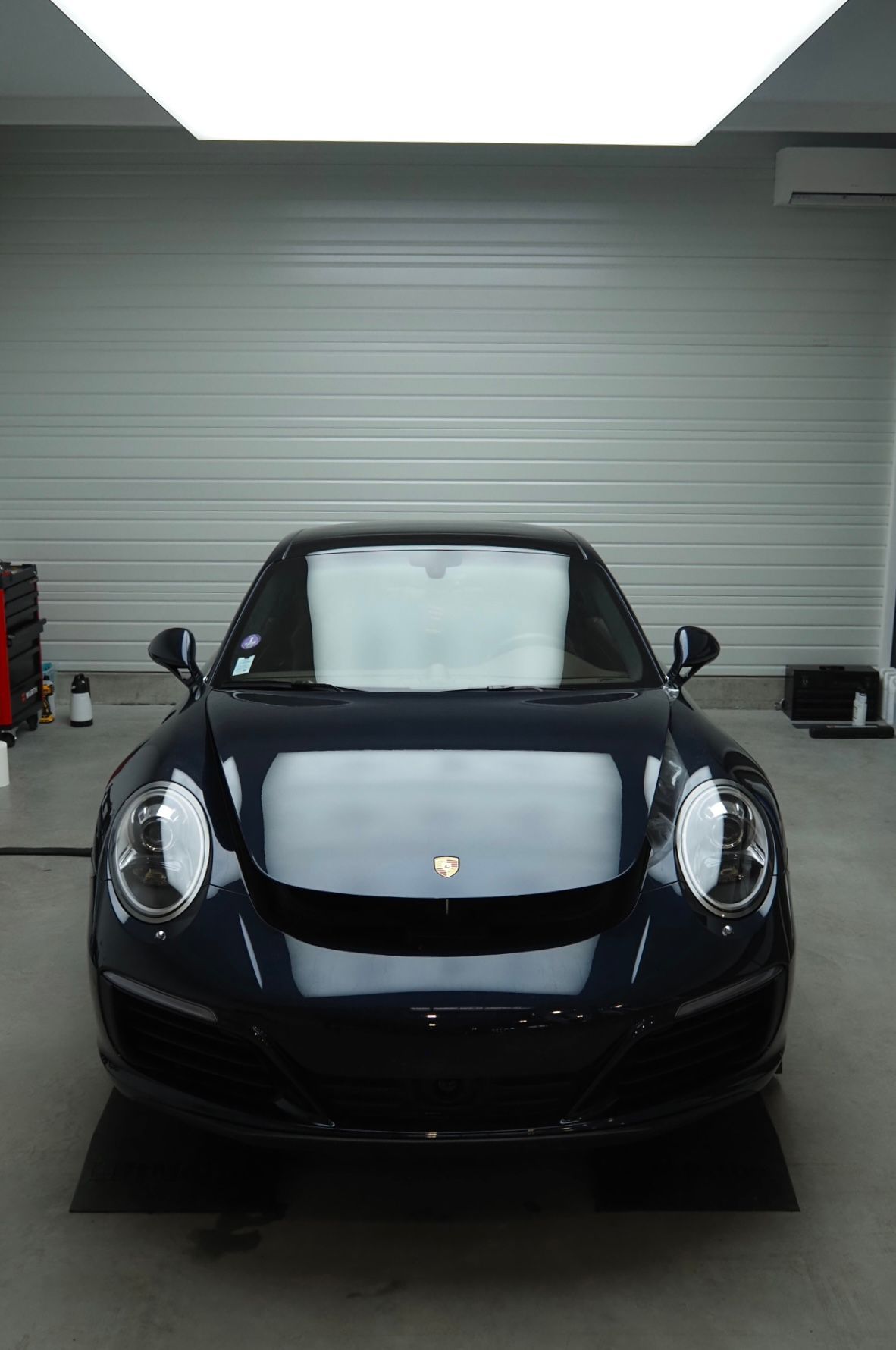 Vue de face d'une Porsche noire dans un garage avec une porte grise fermée derrière.