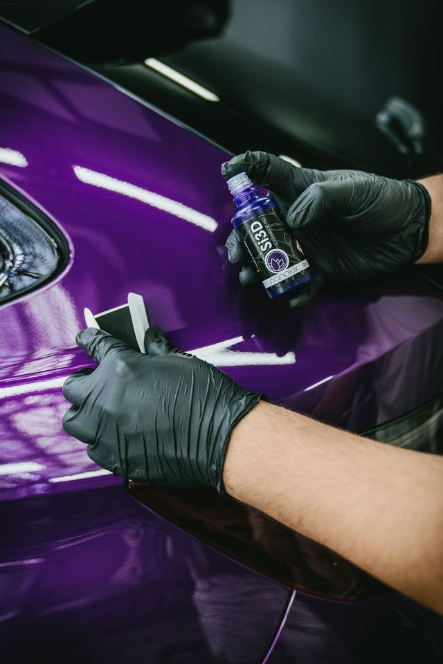 Des mains gantées appliquent un revêtement sur un panneau de voiture violet à l'aide d'un petit flacon et d'un chiffon.