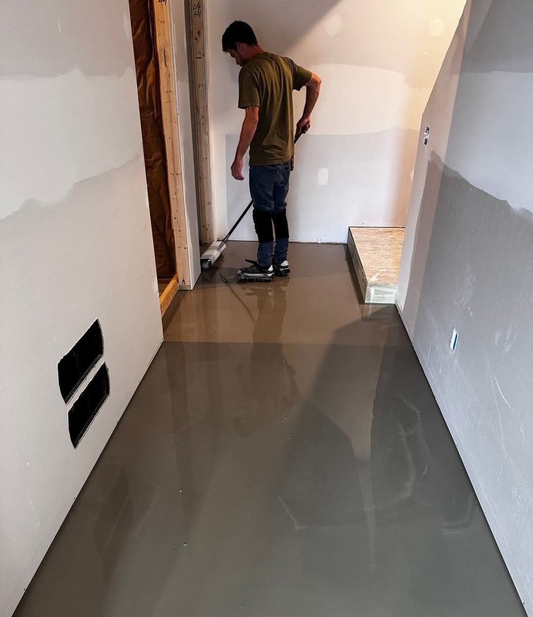 Man leveling wet cement floor with a long-handled tool in a construction area with unfinished walls.