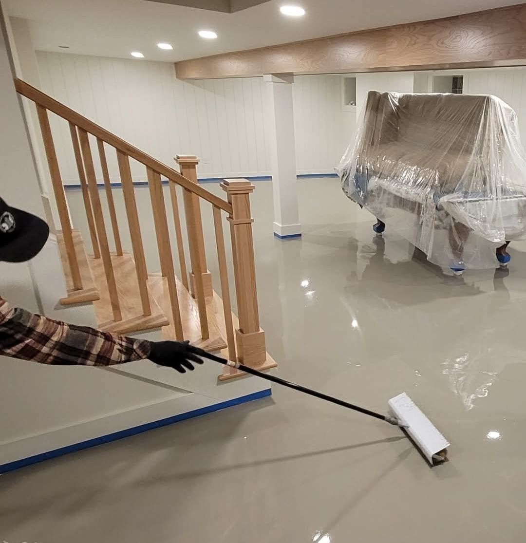 Person applying epoxy coating to a floor in a basement with stairs and a piano covered in plastic.