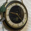 Round brass wall clock with Roman numerals and black face on a stone wall.