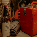 Red Coca-Cola lunch cooler on a rustic shelf with vintage bottles and old objects nearby