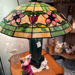 Stained-glass table lamp with floral design on a wooden table beside a figurine and glass dish.
