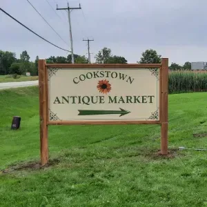 Cookstown Antique Market sign by a roadside on a grassy field, with trees and power lines nearby
