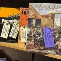 Board game boxes on a shelf, including Napoleon and Pazzle, each marked with handwritten sale notes.