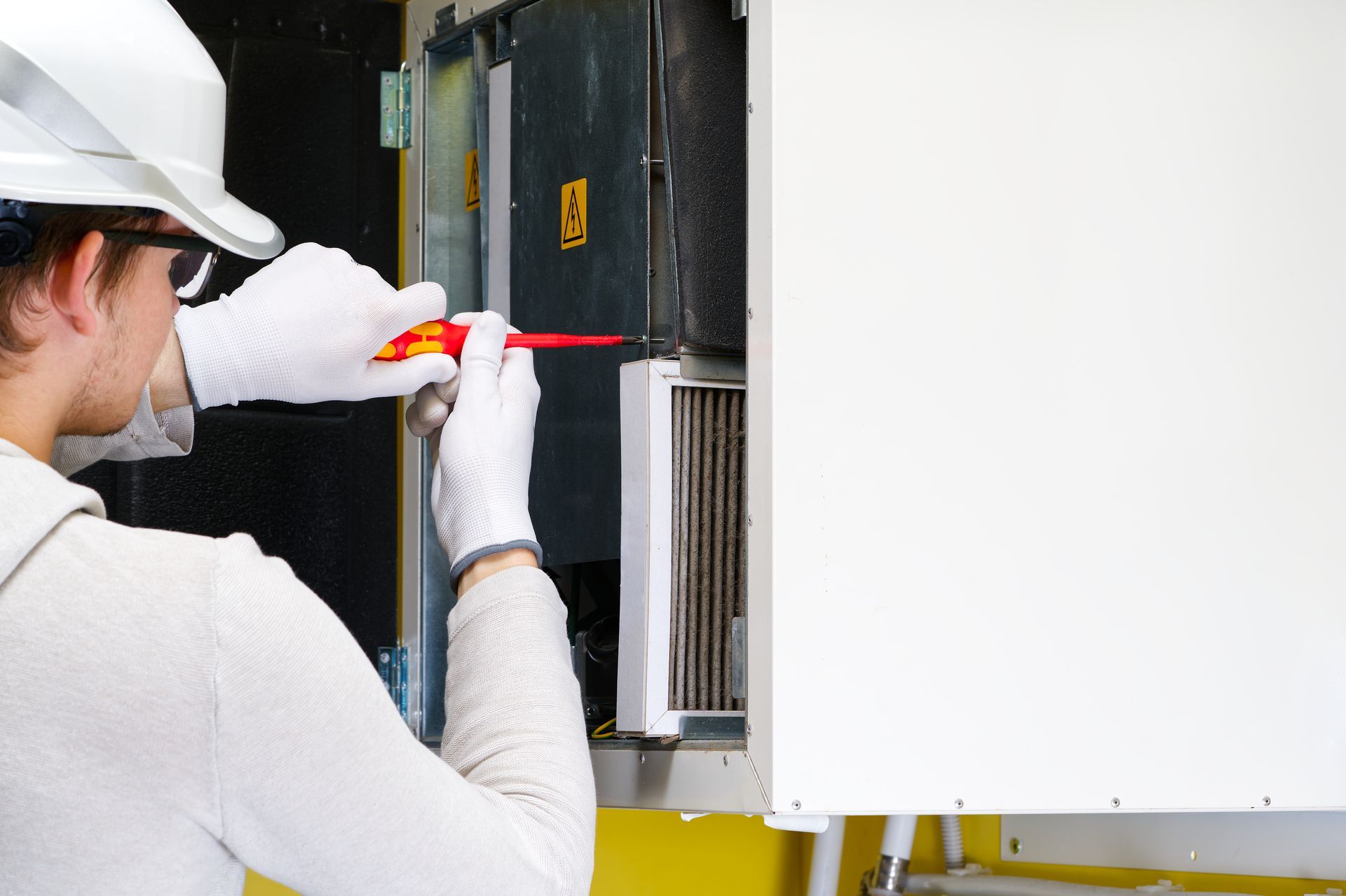 A technician in a white hard hat and gloves uses a screwdriver on equipment.