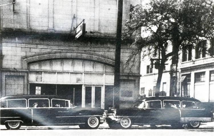 Black-and-white street scene with vintage cars parked in front of a stone building with arched entrance.