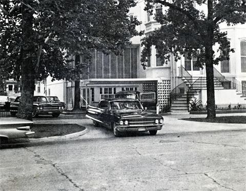 Black-and-white street scene with vintage cars parked beside a building and stairway under tall trees.